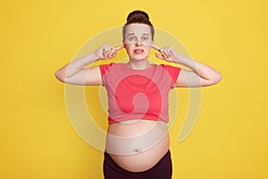 Caucasian expectant mother posing isolated over yellow background, covering her ears with fingers, hears noise or loud music,