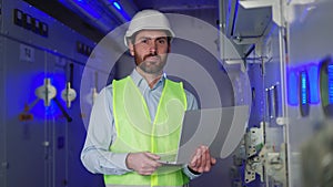 Caucasian factory specialist on laptop in plant with blue bg, smiling at camera.