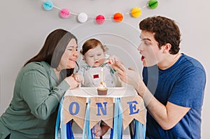 Caucasian family with baby boy celebrating his first birthday at home. Mother father dad with child kid toddler blowing candle on