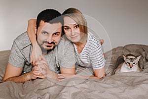 Caucasian family man and woman lying on bed in bedroom at home with oriental cat. Proud smiling pet owners with a domestic animal