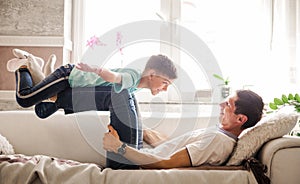 Caucasian family spend time together in living room father lying on couch lift up son, little kid imitates plane imagines himself