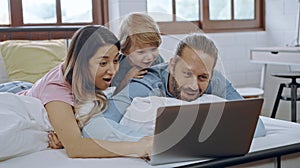 Caucasian family of three using laptop while lying on bed together, browsing internet or watching movie