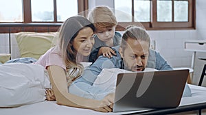 Caucasian family of three using laptop while lying on bed together, browsing internet or watching movie