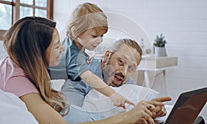 Caucasian family of three using laptop while lying on bed together, browsing internet or watching movie