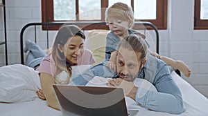 Caucasian family of three using laptop while lying on bed together, browsing internet or watching movie