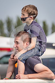 Caucasian father carrying his son on his shoulders outside at an event.  Son has sunglasses under his nose