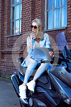 Caucasian Female Biker in Blue Suit Working With Laptop While Sitting on Shiny New Motorcycle  With Cup of Coffee Outdoors