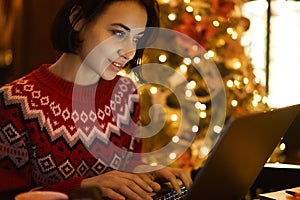 Caucasian female blogger in red winter sweater texting and reading connected to 4g wireless in xmass interior photo