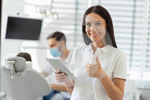 Caucasian female doctor showing thumb up, holding digital tablet and smiling at the camera with colleague working with client on