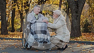 Caucasian female elderly grey-haired woman holding husband hand senior man with disability on wheelchair supporting old