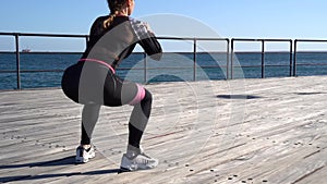Caucasian female fitness model performing banded squats exercise outdoor at sea promenade