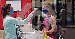 Caucasian female gym receptionist in face mask taking temperature of masked female client