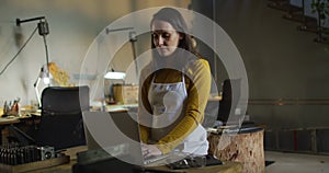 Caucasian female jeweller in workshop wearing apron, sitting at desk, using laptop