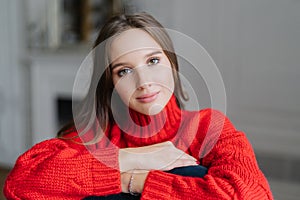 Caucasian female with make up, dark hair, dressd in red warm swater, looks directly at camera with calm expression, poses indoor a