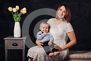 Caucasian female parent and son baby boy sitting together on couch indoors. Happy mother day holiday