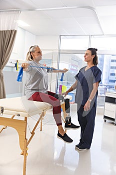 Caucasian female physiotherapist and senior woman with artificial leg using exercise band stretching