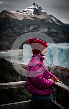 Caucasian female in a pink jacket and hat stands on a railing, admiring the majestic mountain view