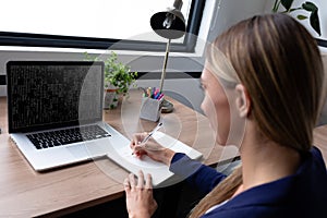 Caucasian female programmer sitting at desk, using laptop with coding on screen, making notes