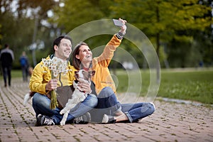 Caucasian female taking selfie with her boyfriend and his dog in a lap, sitting on a path in park