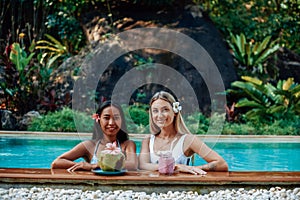 Caucasian female traveller with asian woman in pool with natural juice