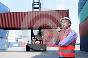 Caucasian foreman man is using walkie talkie to command his workers with crane lifting container in background in container depot