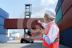 Caucasian foreman man is using walkie talkie to command his workers with crane lifting container in background in container depot