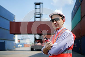 Caucasian foreman man is using walkie talkie to command his workers with crane lifting container in background in container depot