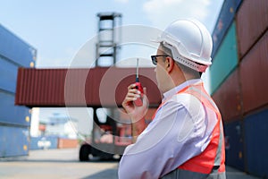 Caucasian foreman man is using walkie talkie to command his workers with crane lifting container in background in container depot