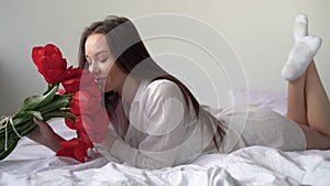 Caucasian girl 20 s in a shirt lies on the bed with a bouquet of flowers, sniffs them and enjoys the morning. Red tulips