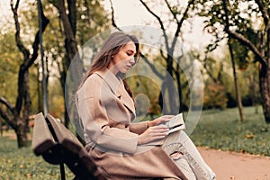 A Caucasian girl in a beige coat and blue jeans sits on a bench in a park in autumn and reads a book