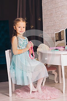 caucasian girl in a blue dress sits on a chair at the dressing table and holds pink beads in her hands