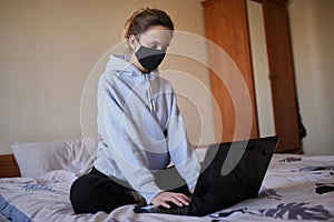 Caucasian girl in blue hoodie and black mask works on laptop at home .
