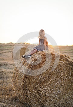 caucasian girl enjoy having fun sitting on top over golden hay bale on wheat harvested field near farm. Happy childhood