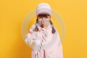 Caucasian girl feeling bed smell, looking smiling at camera and closing nose with finger, little girl wearing rose cap and poncho