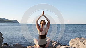 Caucasian girl meditating in a black swimsuit. Woman practices yoga at the view of the ocean or sea at sunset light