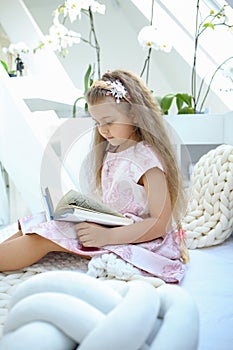 Caucasian girl in a pink dress is reading a book while sitting in a children`s room