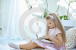 Caucasian girl in a pink dress is reading a book while sitting in a children`s room