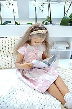 Caucasian girl in a pink dress reads a book while sitting in a children`s room