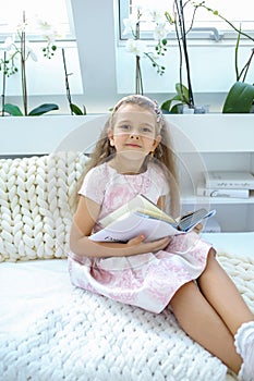 Caucasian girl in a pink dress reads a book while sitting in a children`s room