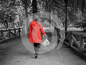 A Caucasian girl with a red gabardine walks through a park with a transparent umbrella in her hand