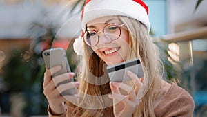 Caucasian girl in santa hat makes internet order using telephone and credit card millennial woman smiling happily buying