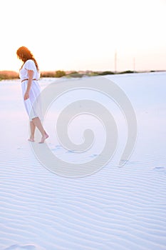 Caucasian girl wearing white dress walking barefoot on sand.