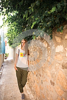 Caucasian girl in white T-shirt with fruits, green pants and gray sneakers is walking along a narrow ancient street with trees and