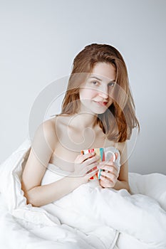 Caucasian girl woman with long red hair sitting in bed wrapped covered with blanket, holding cup