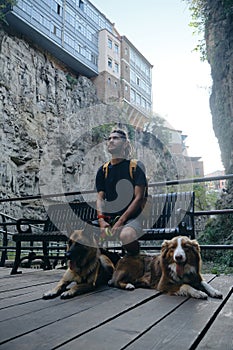 Caucasian guy with dreadlocks is sitting on bench in center of old town with two dogs and enjoying architecture