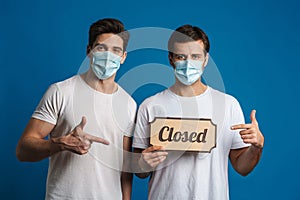 Caucasian guys in masks gesturing while showing closed sign board