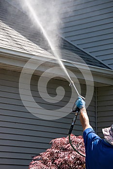 A caucasian hand with a latex glove arm holding a water sprayer wand power washing the gutters and roof of a house