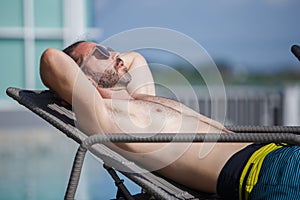 Caucasian handsome man wearing sunglasses lying at near swimming pool, weekend relax luxury travel poolside. Summer vacation