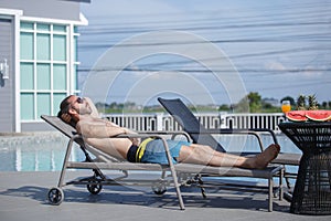 Caucasian handsome man wearing sunglasses lying at near swimming pool, weekend relax luxury travel poolside. Summer vacation