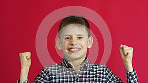 Caucasian happy boy 7-8 years old victoriously waving his hands says yes looking at the camera. Studio shooting on a red backgroun